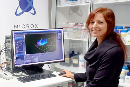 Alenka Lovy smiling in a lab, posing in front of a computer with imaging software open next to a light-sheet microscope.